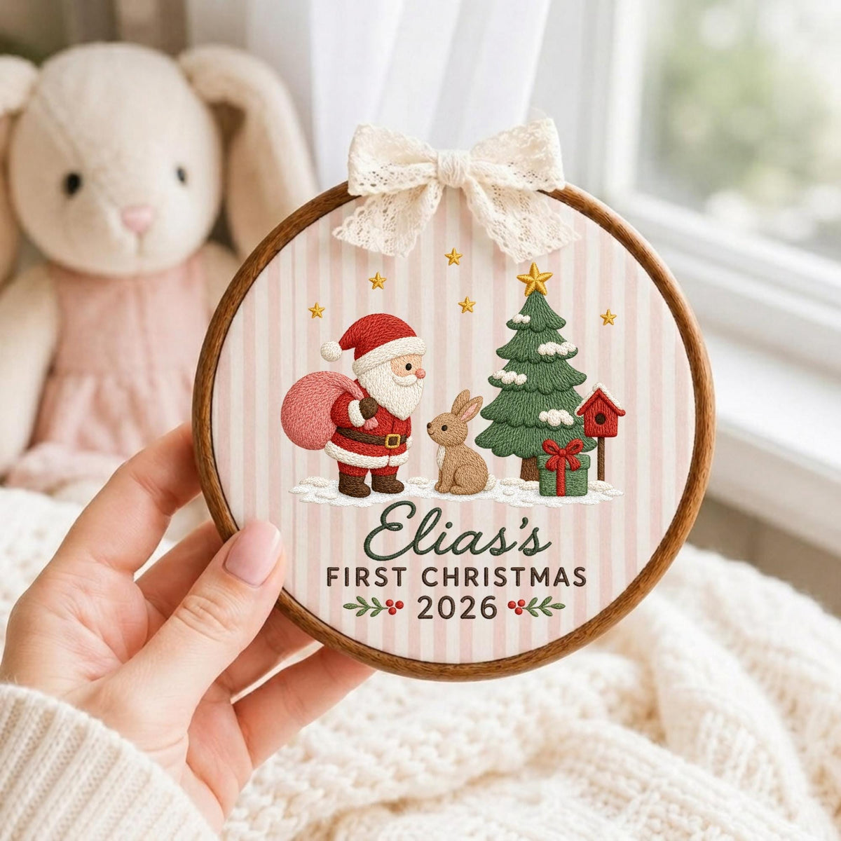 May include: A round wooden embroidery hoop ornament featuring a light pink and white striped fabric background. The design includes an embroidered Santa Claus, a rabbit, a Christmas tree, and a birdhouse. Text reads: Elias's FIRST CHRISTMAS 2026.