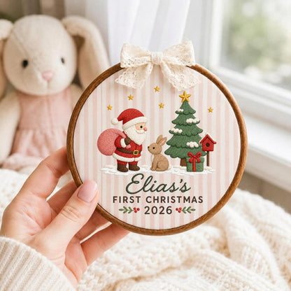 May include: A round wooden embroidery hoop ornament featuring a light pink and white striped fabric background. The design includes an embroidered Santa Claus, a rabbit, a Christmas tree, and a birdhouse. Text reads: Elias's FIRST CHRISTMAS 2026.