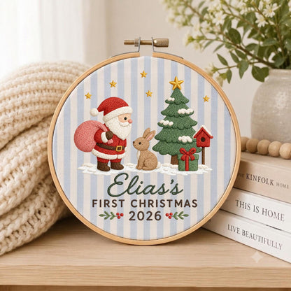 May include: A circular embroidery hoop featuring a light blue and white striped fabric background. The design includes an embroidered Santa Claus, a brown rabbit, a green Christmas tree, and a red birdhouse. Text reads: Elia's FIRST CHRISTMAS 2026.