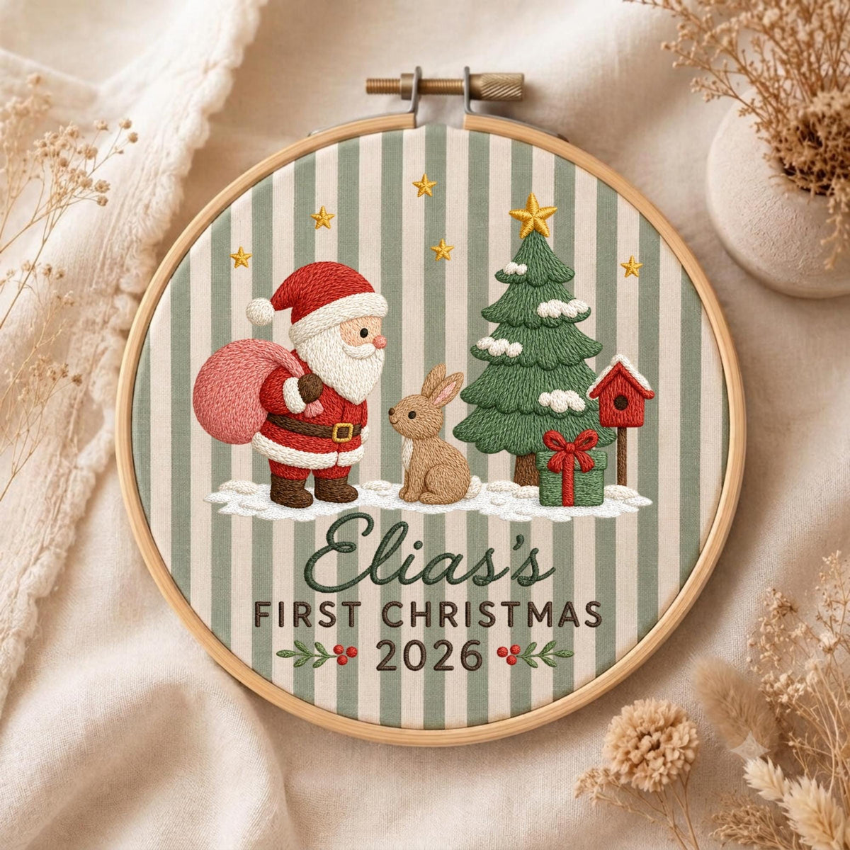 May include: A circular embroidery hoop featuring a striped fabric background with a stitched scene of Santa Claus, a brown rabbit, a green Christmas tree, and a red birdhouse. The text reads Elias's FIRST CHRISTMAS 2026 with small holly accents.