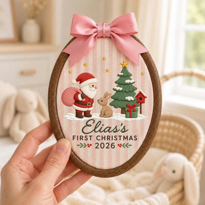 May include: An oval wooden embroidery hoop ornament featuring a pink and white striped fabric background. It displays an embroidered Santa Claus, a brown rabbit, a green Christmas tree, and a red birdhouse. Text reads: Elias's FIRST CHRISTMAS 2026.