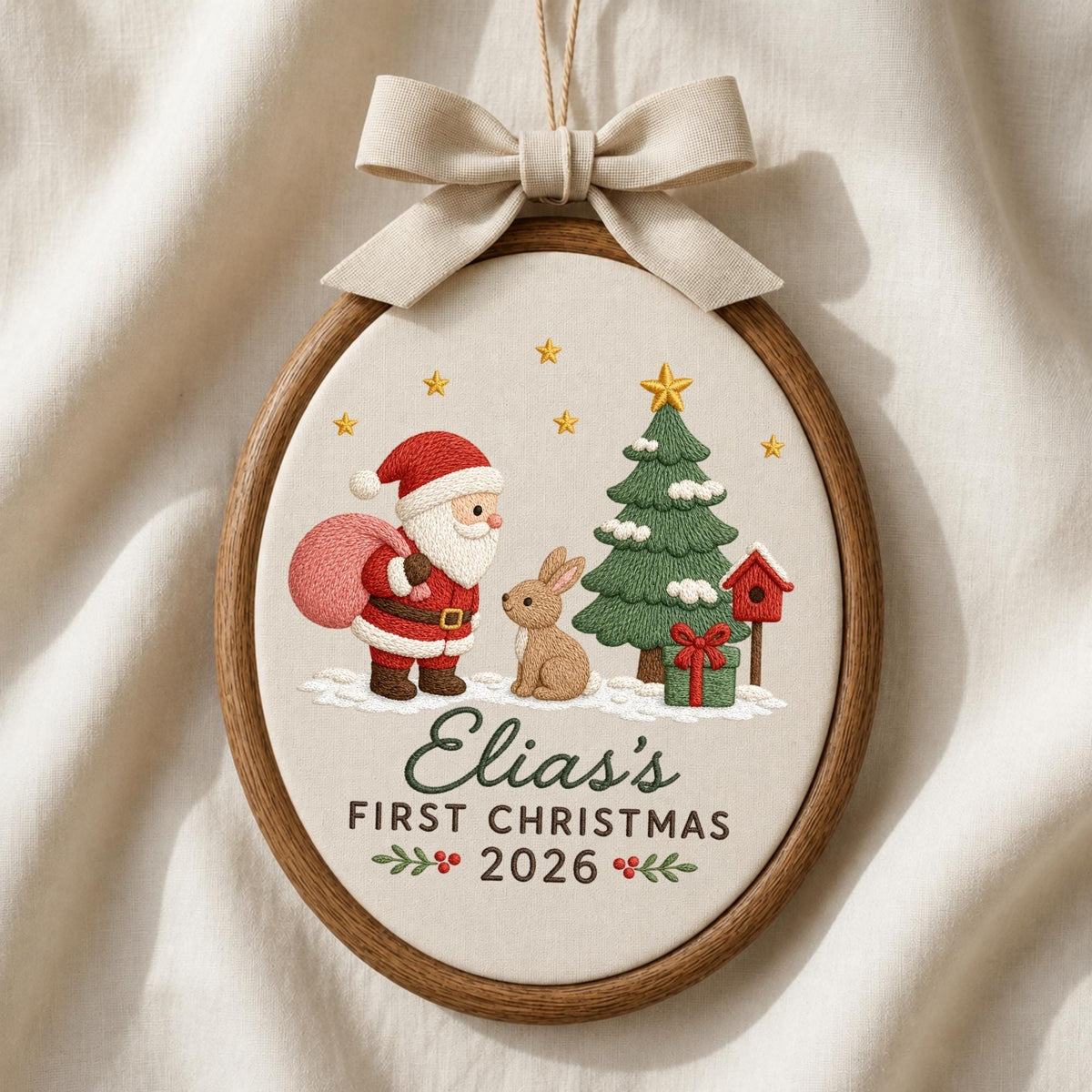 May include: An oval wooden embroidery hoop featuring a cream fabric background with a stitched design of Santa Claus, a brown rabbit, a green Christmas tree, and a red birdhouse. The text reads Elias's FIRST CHRISTMAS 2026 with a beige bow on top.
