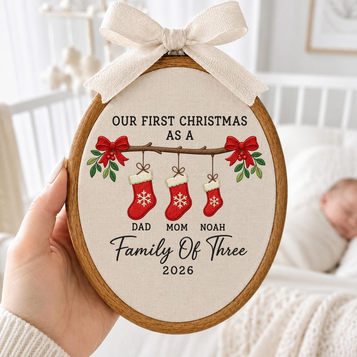 May include: An oval wooden embroidery hoop featuring a cream fabric background with a beige ribbon bow at the top. The design shows three red stockings hanging from a branch with red bows and green leaves, with text reading OUR FIRST CHRISTMAS AS A FAMILY OF THREE DAD MOM NOAH 2026.