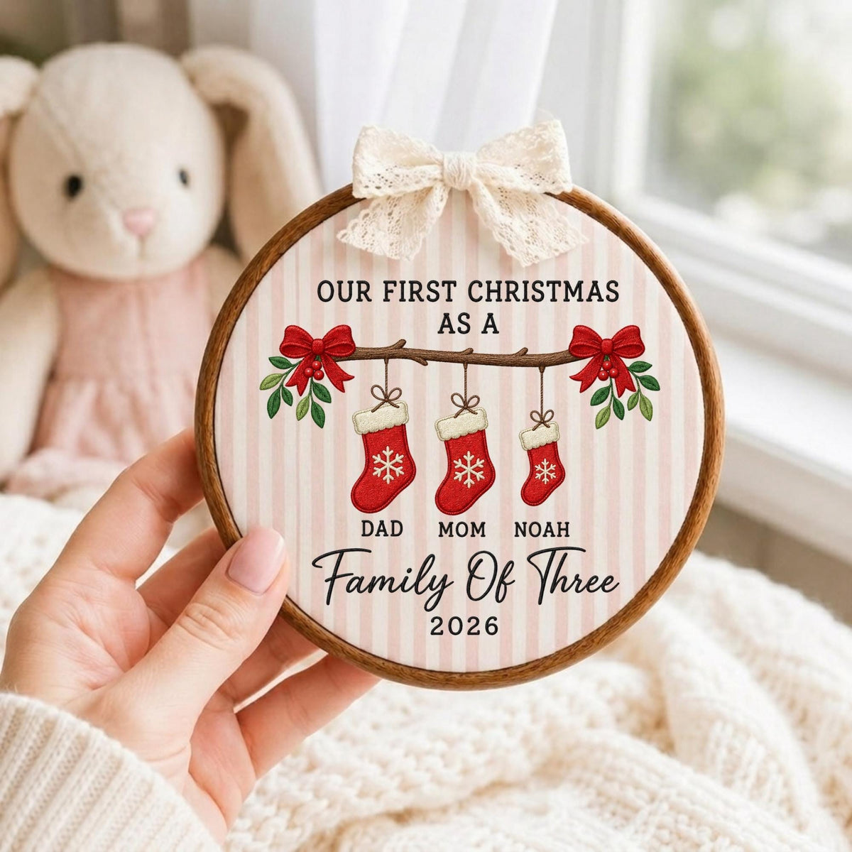 May include: A round wooden embroidery hoop ornament featuring a light pink and white striped background. It displays the text OUR FIRST CHRISTMAS AS A FAMILY OF THREE 2026, with three red stockings labeled DAD, MOM, and NOAH hanging from a branch.