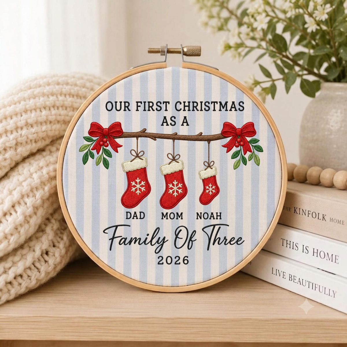 May include: A circular embroidery hoop featuring a light blue and white striped fabric background. The design includes a brown branch with three red Christmas stockings hanging from it, each with a white snowflake, labeled DAD, MOM, and NOAH.