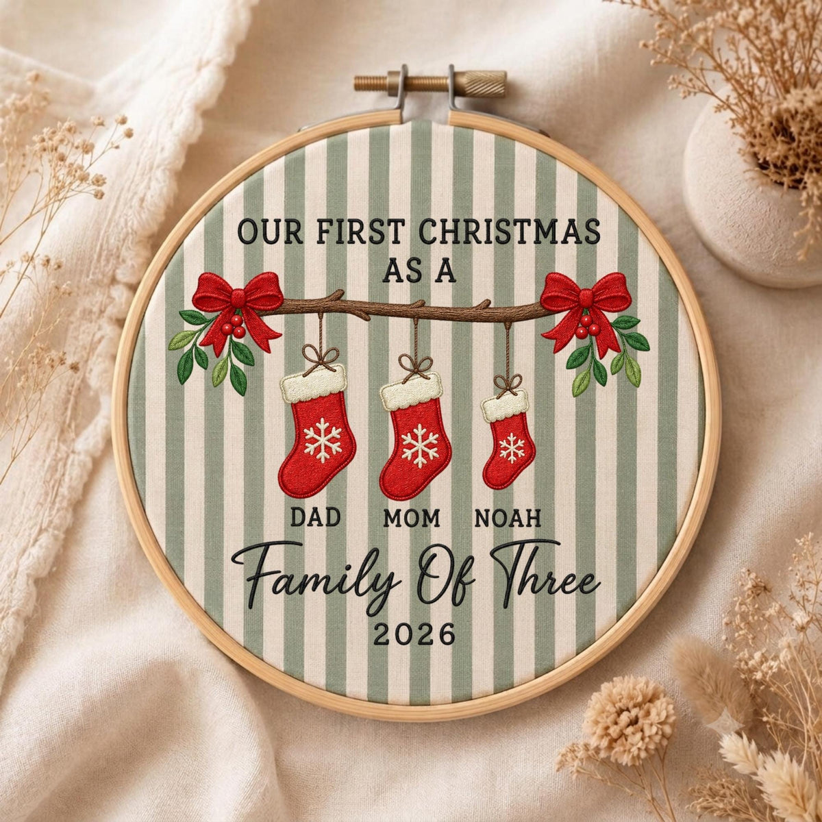 May include: A circular embroidery hoop featuring a light green and white striped fabric with the text "OUR FIRST CHRISTMAS AS A" above a brown branch holding three red stockings with white snowflakes, labeled "DAD", "MOM", "NOAH", "Family Of Three", and "2026".