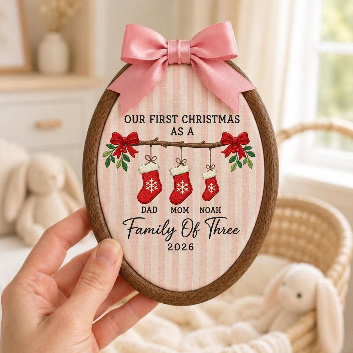 May include: An oval wooden embroidery hoop frame containing fabric with pink vertical stripes. The text reads OUR FIRST CHRISTMAS AS A FAMILY OF THREE 2026. Three red stockings hang from a branch with the names DAD, MOM, and NOAH. A pink satin bow is attached to the top.