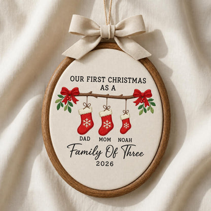 May include: An oval wooden embroidery hoop ornament featuring cream fabric with the text "OUR FIRST CHRISTMAS AS A Family Of Three 2026". Below the text, three red stockings with white cuffs and snowflake designs hang from a branch with red bows.