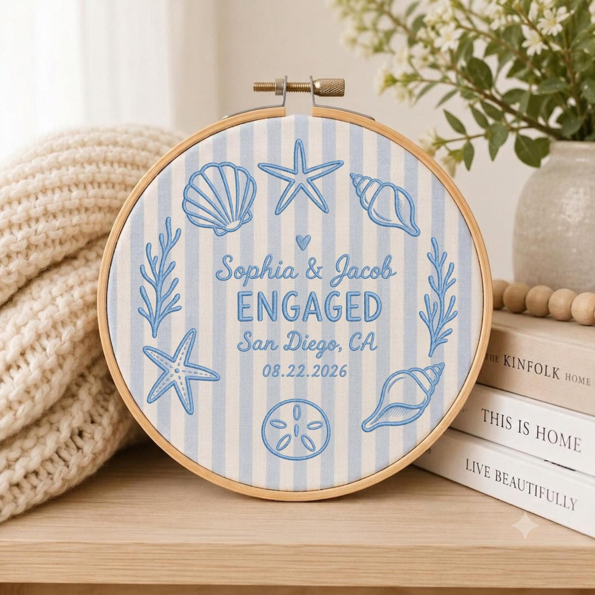 May include: A circular embroidery hoop featuring light blue and white vertical stripes with blue embroidered sea life motifs including seashells, starfish, and seaweed. The center text reads Sophia &amp; Jacob ENGAGED San Diego, CA 08.22.2026.