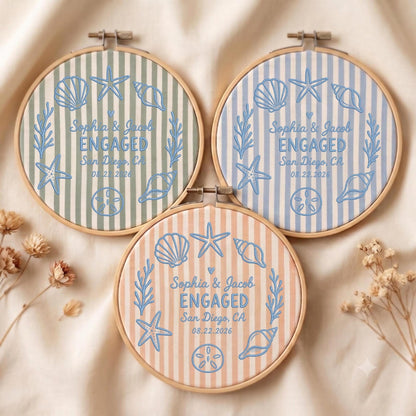 May include: Three circular wooden embroidery hoops featuring striped fabric in sage green, light blue, and peach. Each hoop displays blue embroidered sea life motifs, including seashells and starfish, with the text Sophia &amp; Jacob ENGAGED San Diego, CA 08.22.2026.