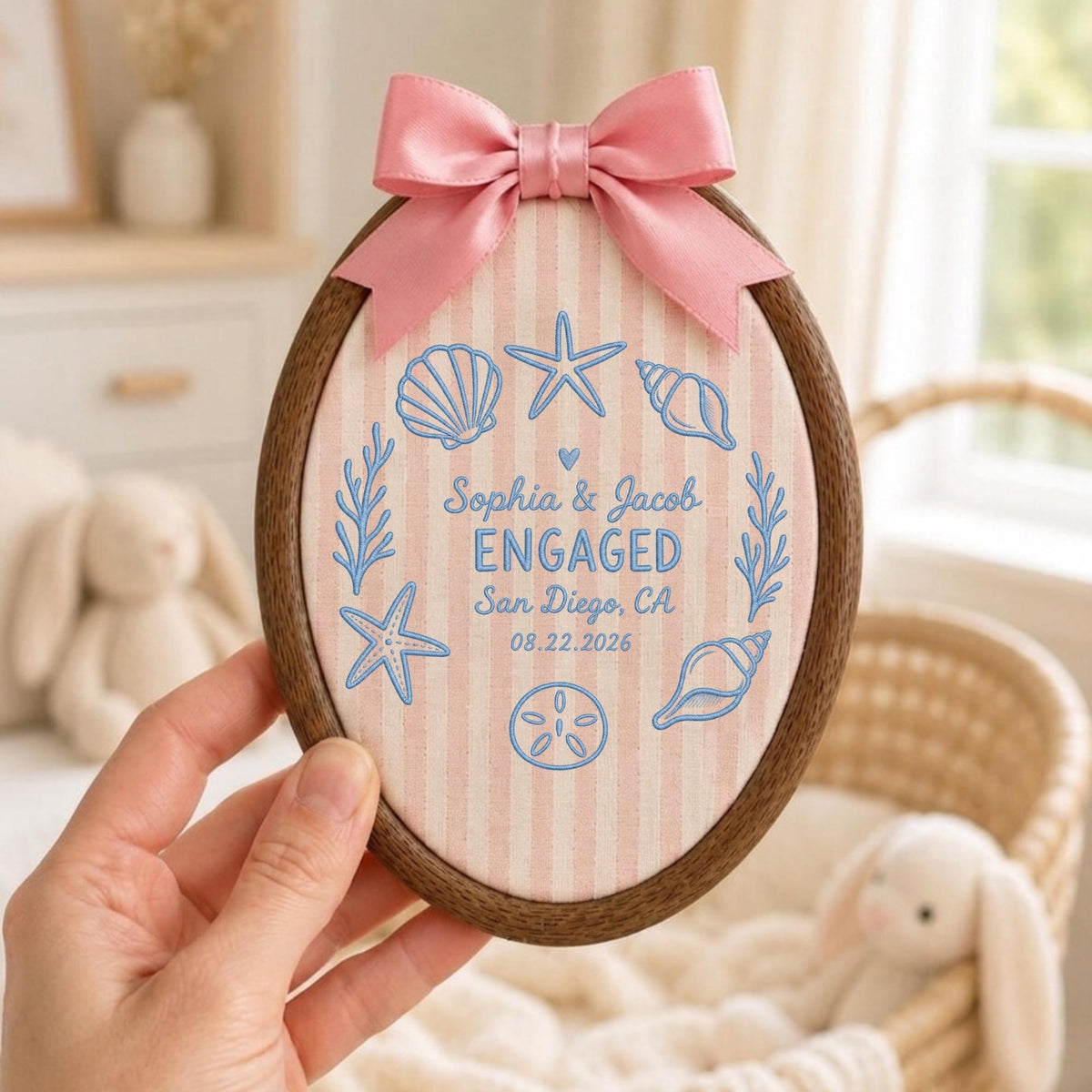 May include: An oval wooden embroidery hoop frame containing fabric with light pink vertical stripes. The fabric features blue embroidered seashells, starfish, and the text: Sophia &amp; Jacob ENGAGED San Diego, CA 08.22.2026. A pink satin bow is attached to the top.