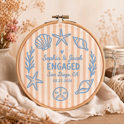 May include: A round wooden embroidery hoop containing fabric with vertical beige and white stripes. The fabric features blue embroidered sea life, including starfish and shells, and the text: Sophia &amp; Jacob ENGAGED San Diego, CA 08.22.2026.