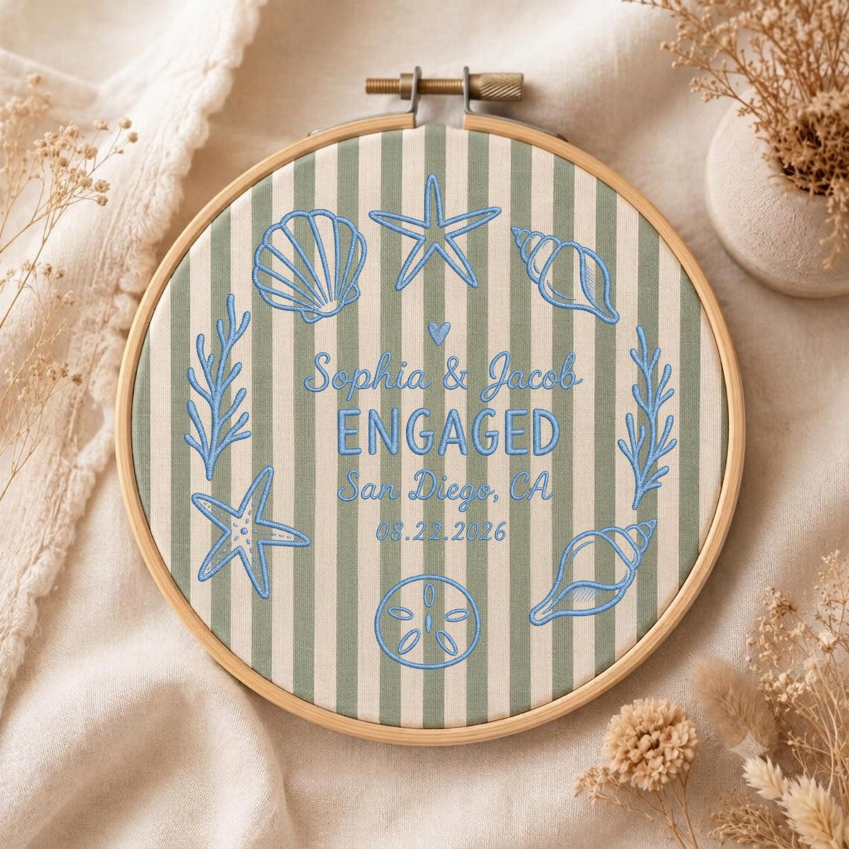 May include: A round wooden embroidery hoop containing fabric with vertical sage green and white stripes. The fabric features blue embroidered sea-themed motifs including seashells, starfish, and coral, with the text Sophia &amp; Jacob ENGAGED San Diego, CA 08.22.2026.