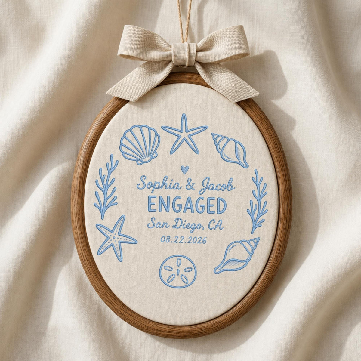 May include: An oval wooden embroidery hoop containing off-white fabric with blue embroidered sea life, including seashells, starfish, and coral. The text reads: Sophia &amp; Jacob ENGAGED San Diego, CA 08.22.2026. A beige fabric bow is attached at the top.