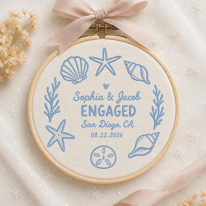 May include: A circular wooden embroidery hoop containing off-white fabric with light blue thread embroidery. The design features seashells, starfish, and seaweed surrounding the text: Sophia &amp; Jacob ENGAGED San Diego, CA 08.22.2026.