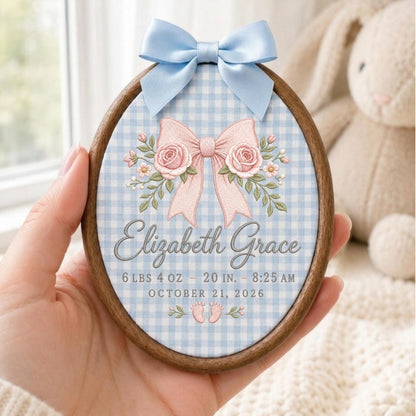May include: An oval wooden embroidery hoop frame containing fabric with a light blue gingham pattern. It features a light blue satin bow at the top, a pink bow with roses, and the text Elizabeth Grace, 6 lbs 4 oz, 20 in, 8:25 am, October 21, 2026.
