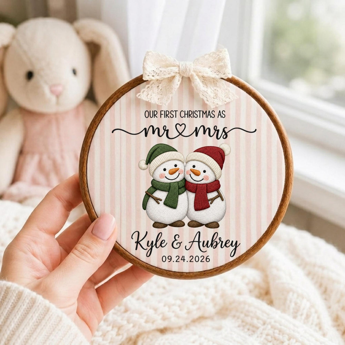May include: A round wooden embroidery hoop ornament featuring a light pink and white striped fabric background. It displays two snowmen with green and red scarves, the text "OUR FIRST CHRISTMAS AS mr &amp; mrs", "Kyle &amp; Aubrey", and "09.24.2026", topped with a cream lace bow.