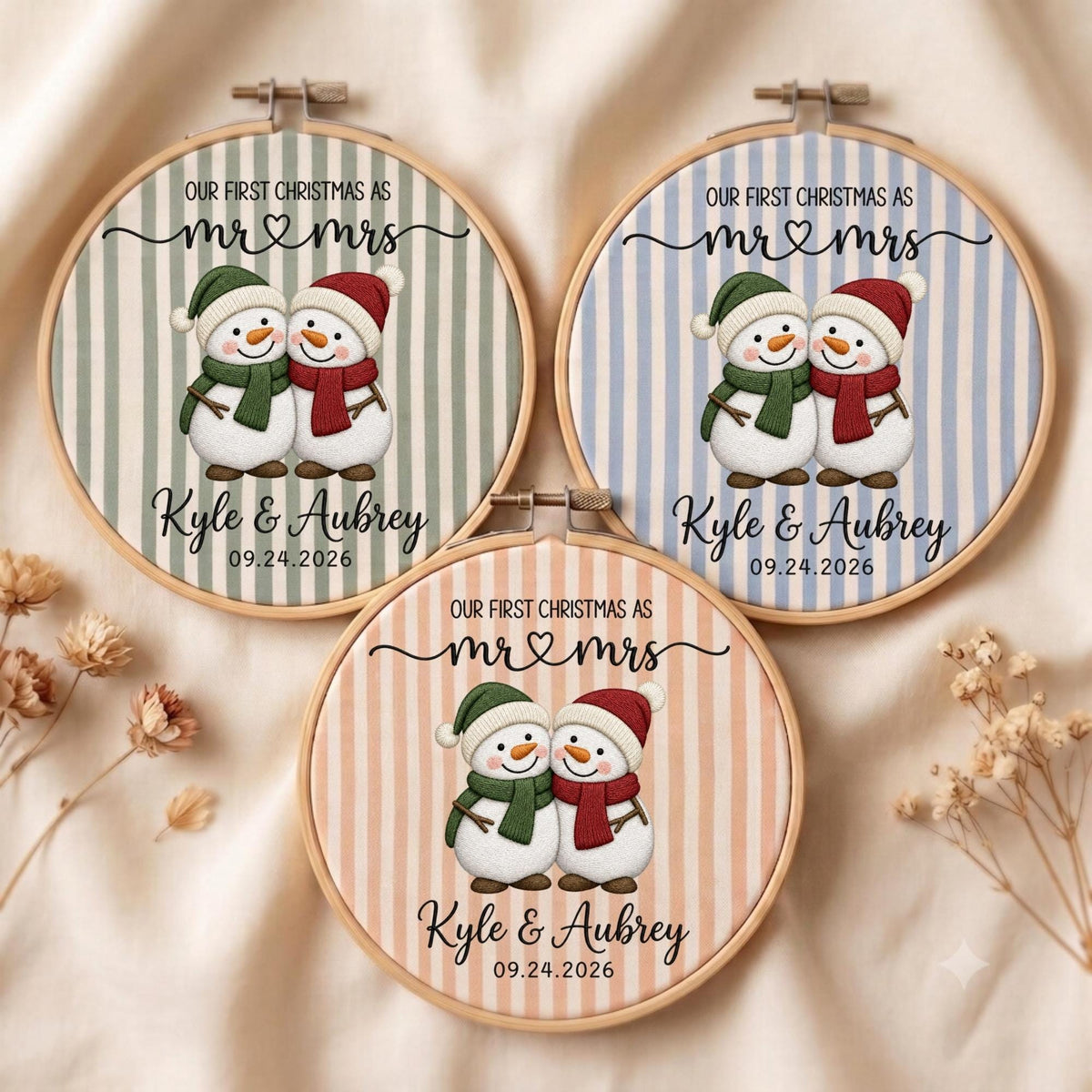 May include: Three circular wooden embroidery hoops featuring fabric with vertical stripes in sage green, light blue, and peach. Each hoop displays two snowmen, the text "OUR FIRST CHRISTMAS AS MR &amp; MRS", the names "Kyle &amp; Aubrey", and "09.24.2026".