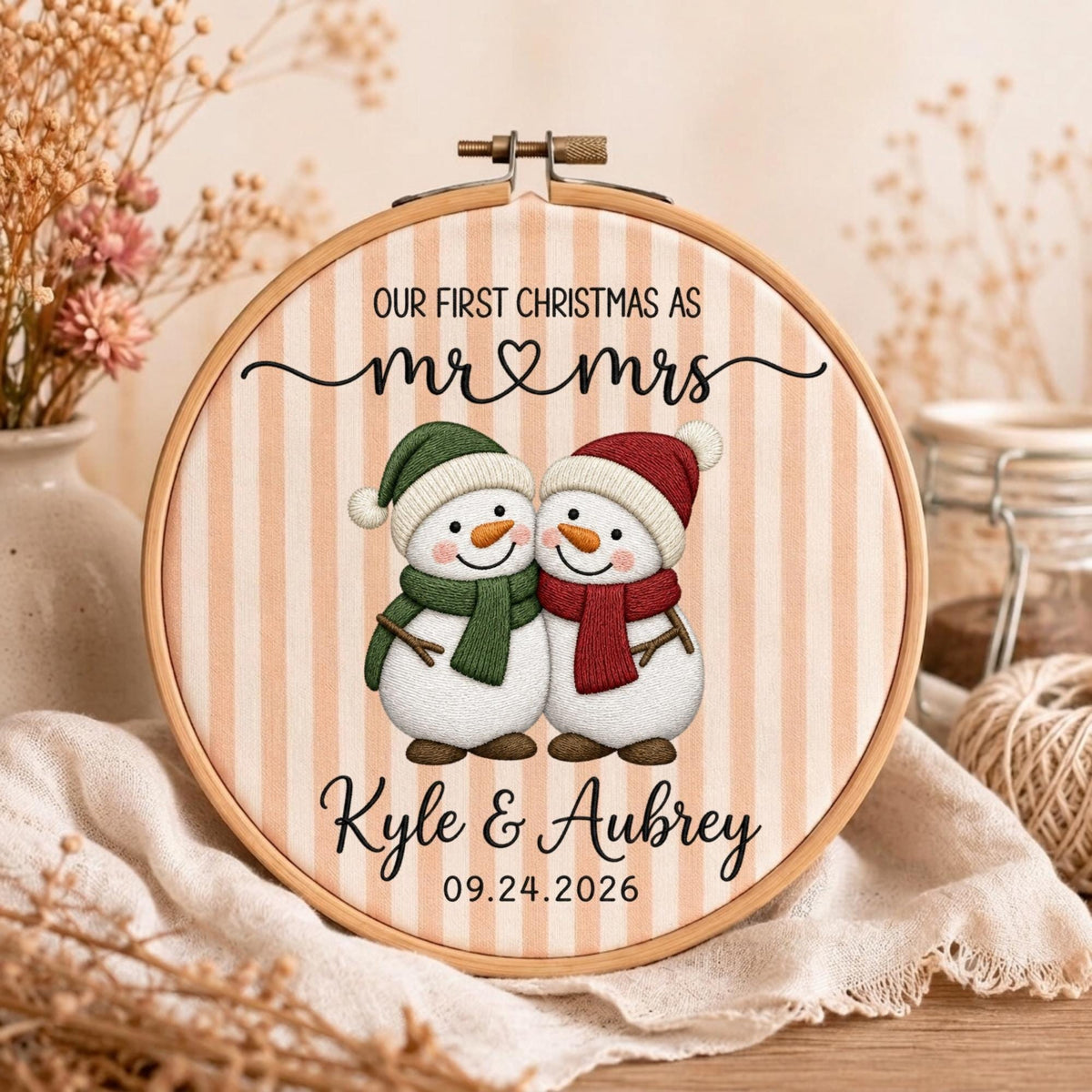 May include: A round wooden embroidery hoop containing fabric with vertical peach stripes. The text reads OUR FIRST CHRISTMAS AS mr &amp; mrs, Kyle &amp; Aubrey, and 09.24.2026. Two snowmen are embroidered in the center, one with a green hat and scarf.