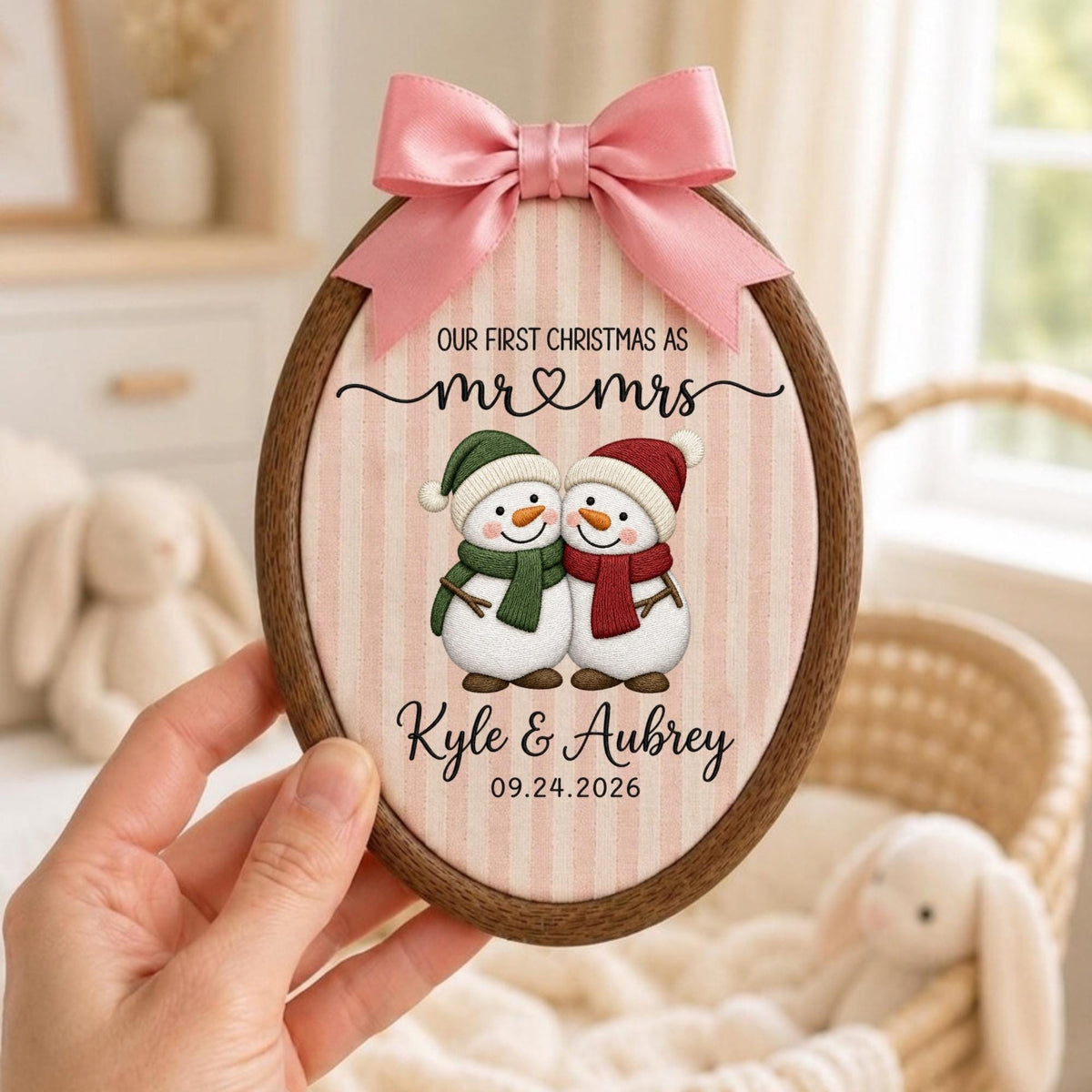May include: An oval wooden embroidery hoop ornament featuring a light pink and white striped fabric background. It displays two snowmen with scarves, the text "OUR FIRST CHRISTMAS AS mr &amp; mrs", "Kyle &amp; Aubrey", and "09.24.2026", topped with a pink satin bow.
