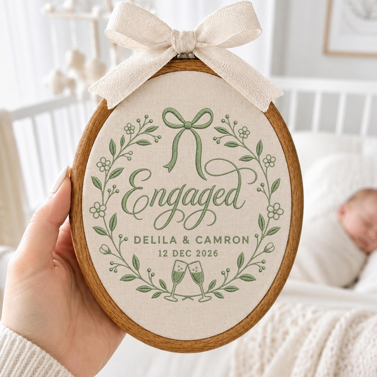 May include: An oval wooden embroidery hoop containing cream-colored fabric with green stitched embroidery. The design features a floral wreath, a bow, the word Engaged, the names Delila &amp; Camron, the date 12 Dec 2026, and two champagne flutes.