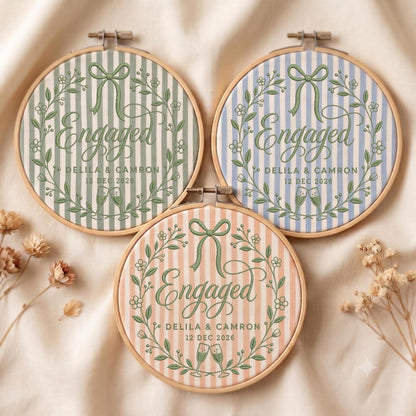 May include: Three circular wooden embroidery hoops featuring striped fabric in sage green, light blue, and peach. Each hoop displays the text Engaged, the names Delila &amp; Camron, and the date 12 Dec 2026, surrounded by a green floral wreath and bow.