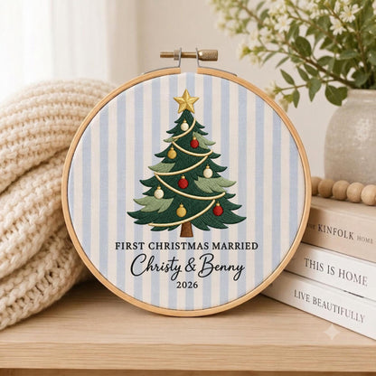 May include: A circular wooden embroidery hoop containing fabric with light blue and white vertical stripes. The center features an embroidered green Christmas tree with a gold star, red and white ornaments, and the text FIRST CHRISTMAS MARRIED Christy &amp; Benny 2026.