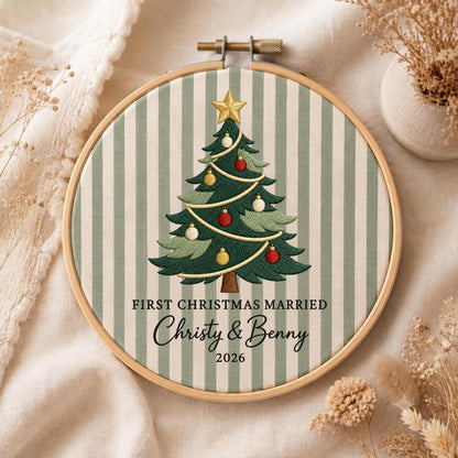May include: A circular wooden embroidery hoop containing fabric with vertical sage green and white stripes. Centered is an embroidered Christmas tree with a gold star, red and gold ornaments, and the text FIRST CHRISTMAS MARRIED Christy &amp; Benny 2026.