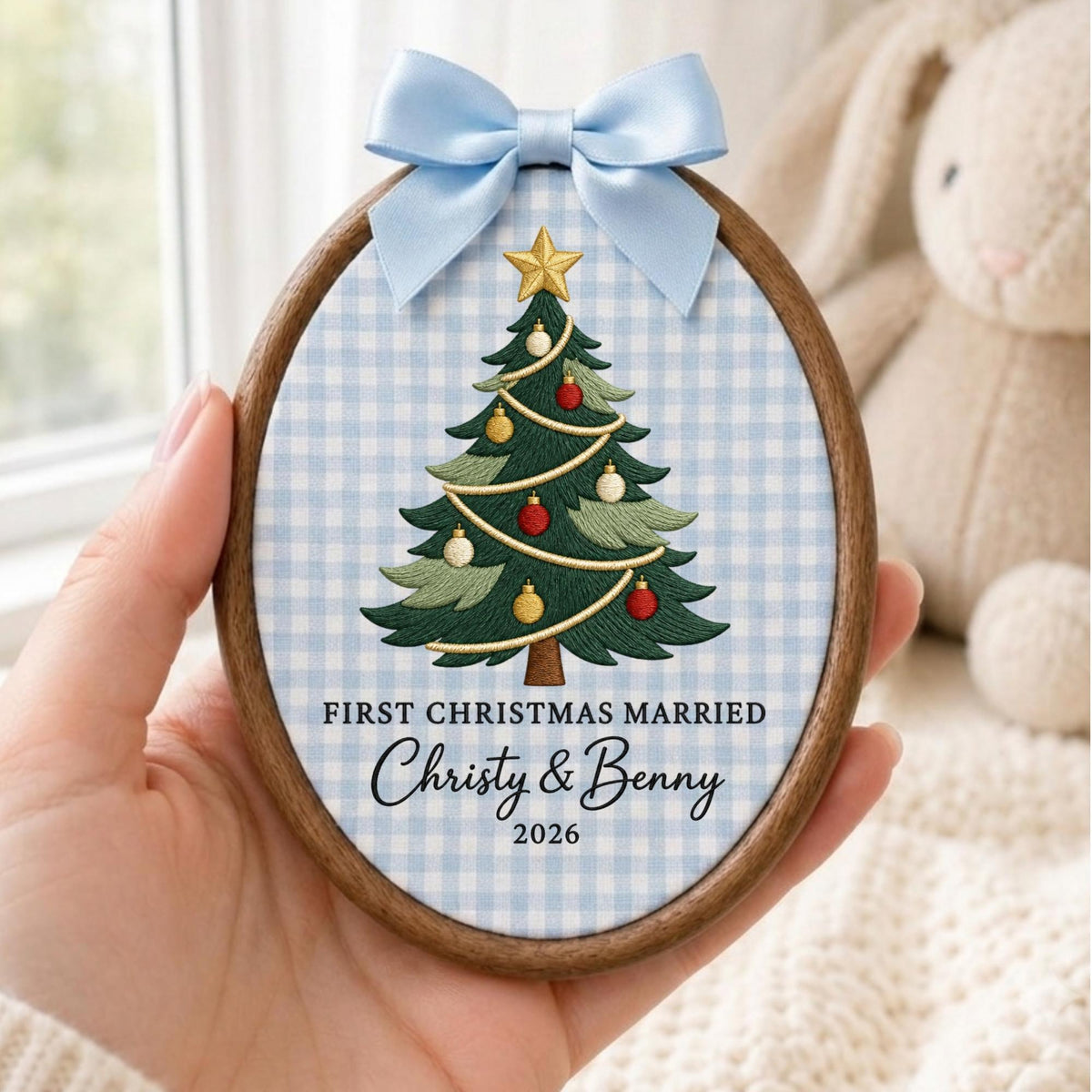 May include: An oval wooden embroidery hoop frame containing fabric with a light blue gingham pattern. The fabric features an embroidered green Christmas tree with red and gold ornaments, a gold star topper, and the text FIRST CHRISTMAS MARRIED Christy &amp; Benny 2026. A light blue satin ribbon bow is attached to the top.