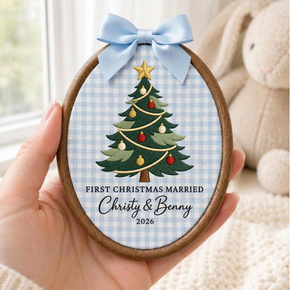 May include: An oval wooden embroidery hoop frame containing fabric with a light blue gingham pattern. The fabric features an embroidered green Christmas tree with red and gold ornaments, a gold star topper, and the text FIRST CHRISTMAS MARRIED Christy &amp; Benny 2026. A light blue satin ribbon bow is attached to the top.