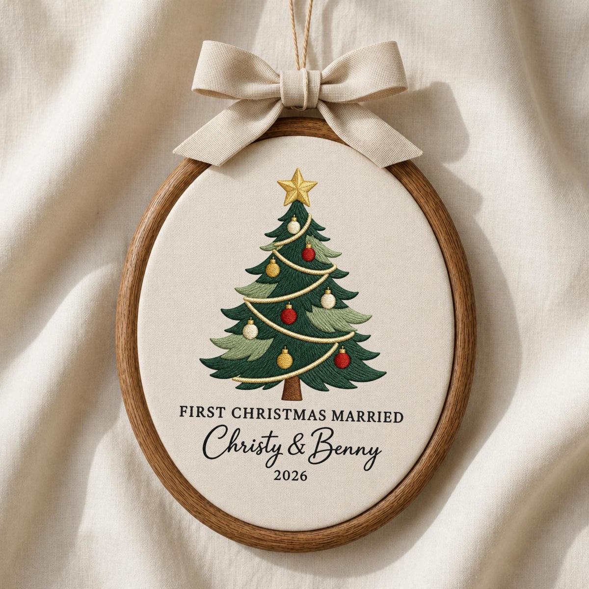 May include: An oval wooden embroidery hoop containing off-white fabric with a stitched Christmas tree, gold star, and ornaments. Text reads: FIRST CHRISTMAS MARRIED, Christy &amp; Benny, 2026. A beige fabric bow is attached to the top of the hoop.