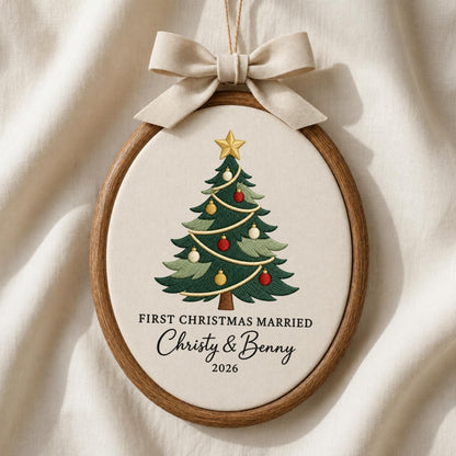 May include: An oval wooden embroidery hoop containing off-white fabric with a stitched Christmas tree, gold star, and ornaments. Text reads: FIRST CHRISTMAS MARRIED, Christy &amp; Benny, 2026. A beige fabric bow is attached to the top of the hoop.