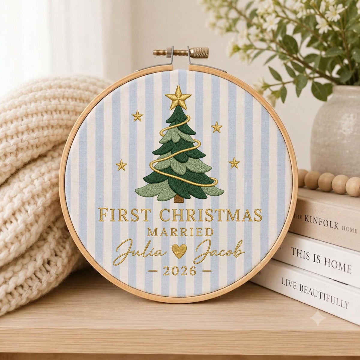May include: A circular wooden embroidery hoop framing light blue and white striped fabric. The center features an embroidered green Christmas tree with a gold star, gold garland, and stars. Text reads: FIRST CHRISTMAS MARRIED Julia and Jacob 2026.