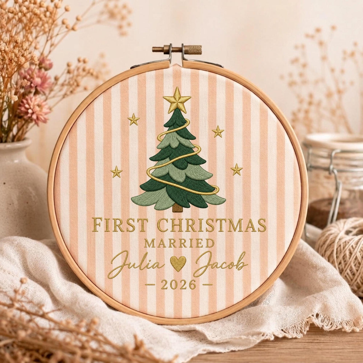May include: A round wooden embroidery hoop containing fabric with vertical light peach stripes. The design features a green Christmas tree with a gold star, gold stars, and the text FIRST CHRISTMAS MARRIED Julia and Jacob 2026.