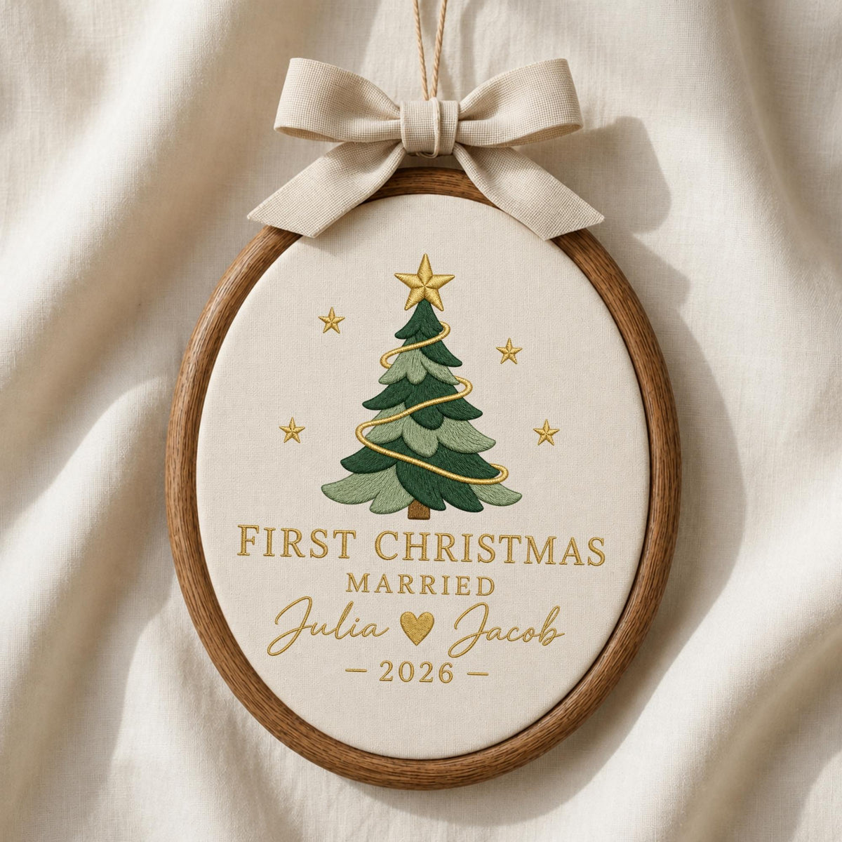 May include: An oval wooden embroidery hoop containing cream fabric with a stitched green Christmas tree, gold stars, and the text "FIRST CHRISTMAS MARRIED Julia &amp; Jacob - 2026 -" in gold thread, topped with a beige fabric bow and hanging string.