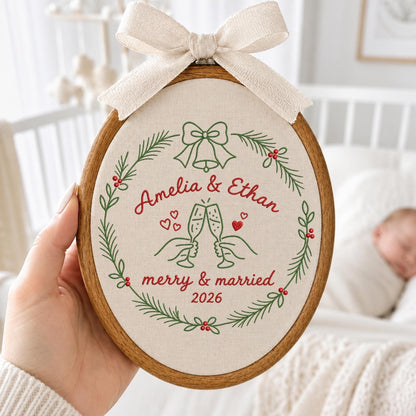 May include: An oval wooden embroidery hoop containing off-white fabric with a green and red stitched wreath design. The embroidery features the text Amelia &amp; Ethan, merry &amp; married, and 2026, with champagne flutes and a bow. A beige ribbon is tied at the top.