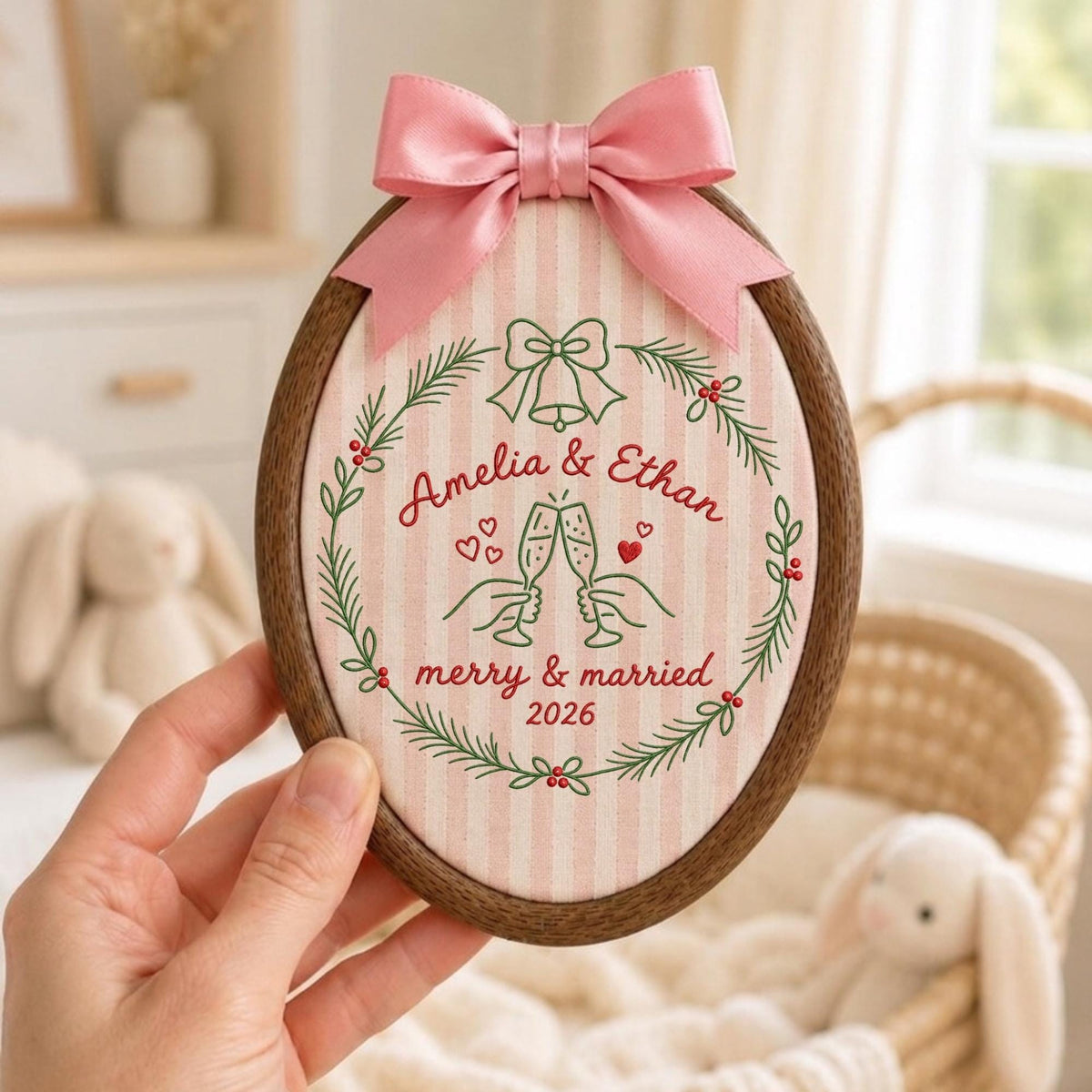 May include: An oval embroidery hoop featuring a light pink and white striped fabric with a dark wood frame. The design includes a green pine wreath, a bell, two hands clinking champagne glasses, and the text Amelia &amp; Ethan merry &amp; married 2026.