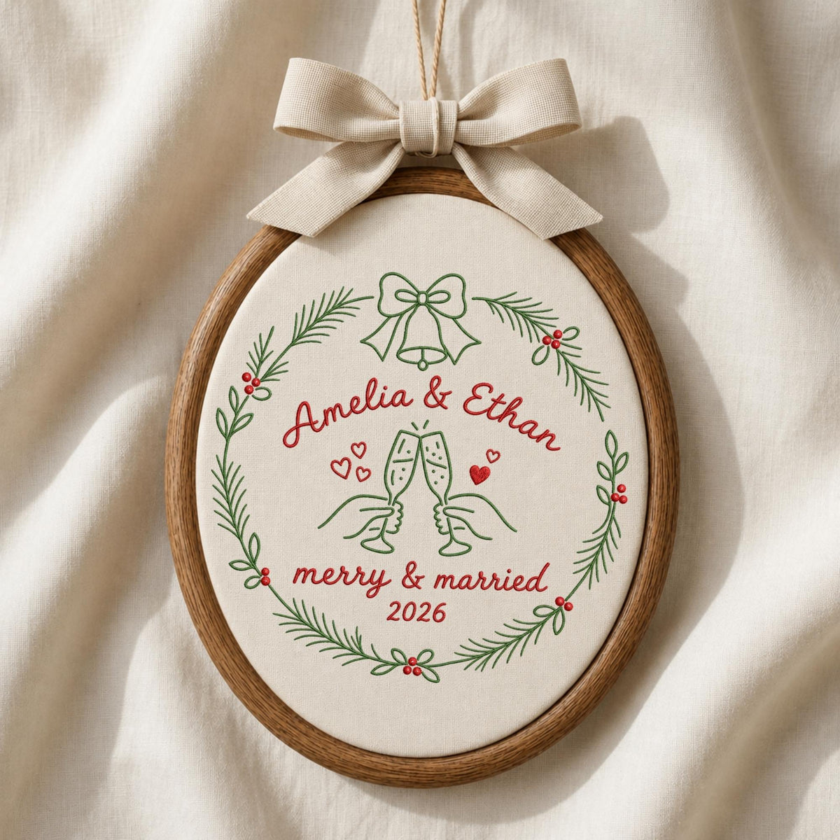 May include: An oval wooden embroidery hoop containing cream fabric with a green pine wreath, red berries, and a beige bow. The embroidery features the text Amelia &amp; Ethan, merry &amp; married 2026, a bell, and two hands clinking champagne glasses.