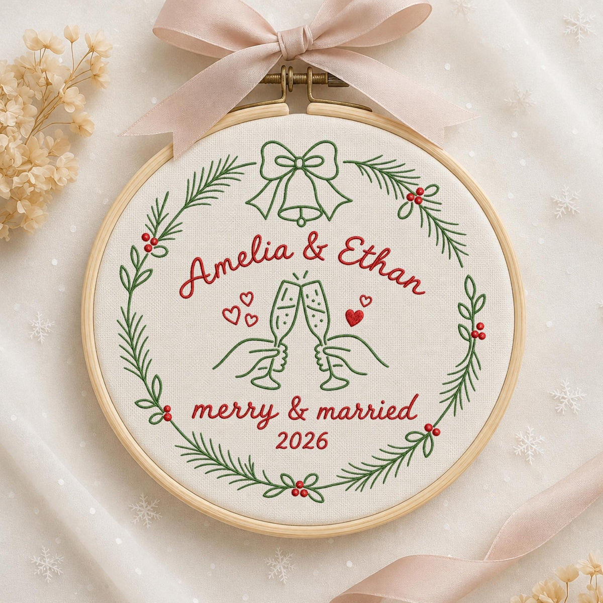 May include: A circular wooden embroidery hoop containing white fabric with green and red stitched designs. The embroidery features a wreath of pine branches, a bell, two hands clinking champagne flutes, and the text Amelia &amp; Ethan merry &amp; married 2026. A light pink ribbon is tied at the top of the hoop.