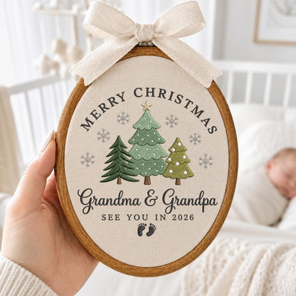 May include: An oval embroidery hoop featuring a beige fabric background with a cream-colored ribbon bow at the top. The design includes three embroidered green Christmas trees, grey snowflakes, and the text Merry Christmas, Grandma &amp; Grandpa, SEE YOU IN 2026, and two small baby footprints.