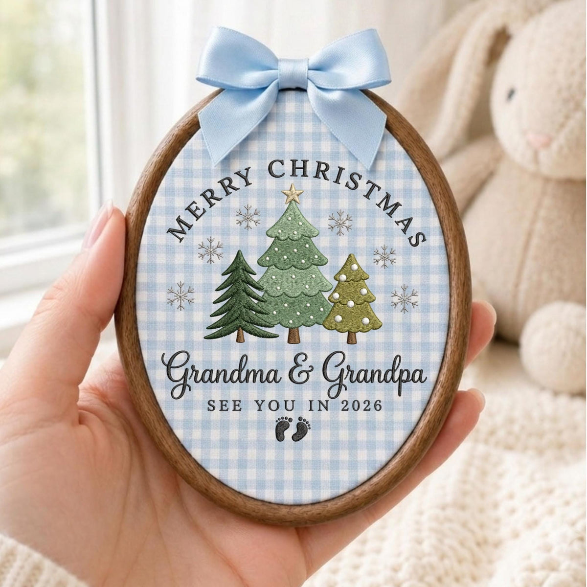 May include: An oval wooden embroidery hoop ornament featuring a light blue and white gingham fabric background. It displays three embroidered Christmas trees, snowflakes, and the text Merry Christmas, Grandma &amp; Grandpa, SEE YOU IN 2026, with baby footprints and a light blue satin bow.