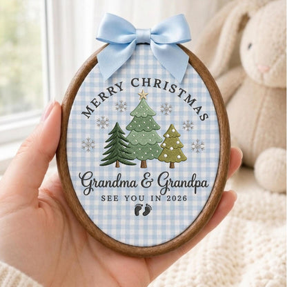 May include: An oval wooden embroidery hoop ornament featuring a light blue and white gingham fabric background. It displays three embroidered Christmas trees, snowflakes, and the text Merry Christmas, Grandma &amp; Grandpa, SEE YOU IN 2026, with baby footprints and a light blue satin bow.