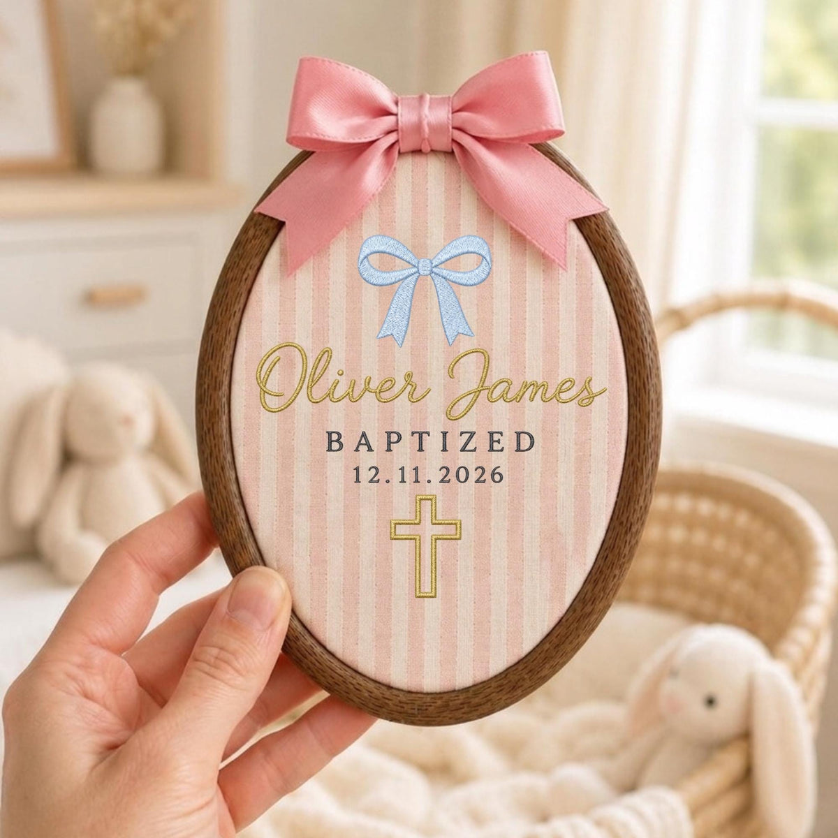 May include: An oval wooden embroidery hoop containing fabric with pink vertical stripes. A pink satin bow is attached to the top. The fabric features a light blue bow graphic, the name Oliver James, the word BAPTIZED, the date 12.11.2026, and a gold cross.