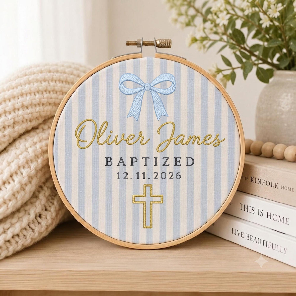 May include: A circular wooden embroidery hoop framing fabric with light blue and white vertical stripes. The design features a light blue bow, the gold embroidered text Oliver James, the word BAPTIZED, the date 12.11.2026, and a gold cross.