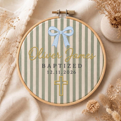 May include: A circular wooden embroidery hoop containing fabric with sage green and white vertical stripes. It features an embroidered light blue bow, the gold text Oliver James, the black text BAPTIZED 12.11.2026, and a gold cross.