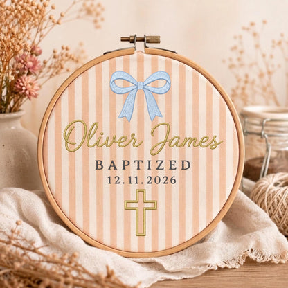 May include: A circular wooden embroidery hoop framing fabric with vertical peach and white stripes. The design features a light blue bow, the name Oliver James in gold script, the text BAPTIZED 12.11.2026, and a gold embroidered cross.