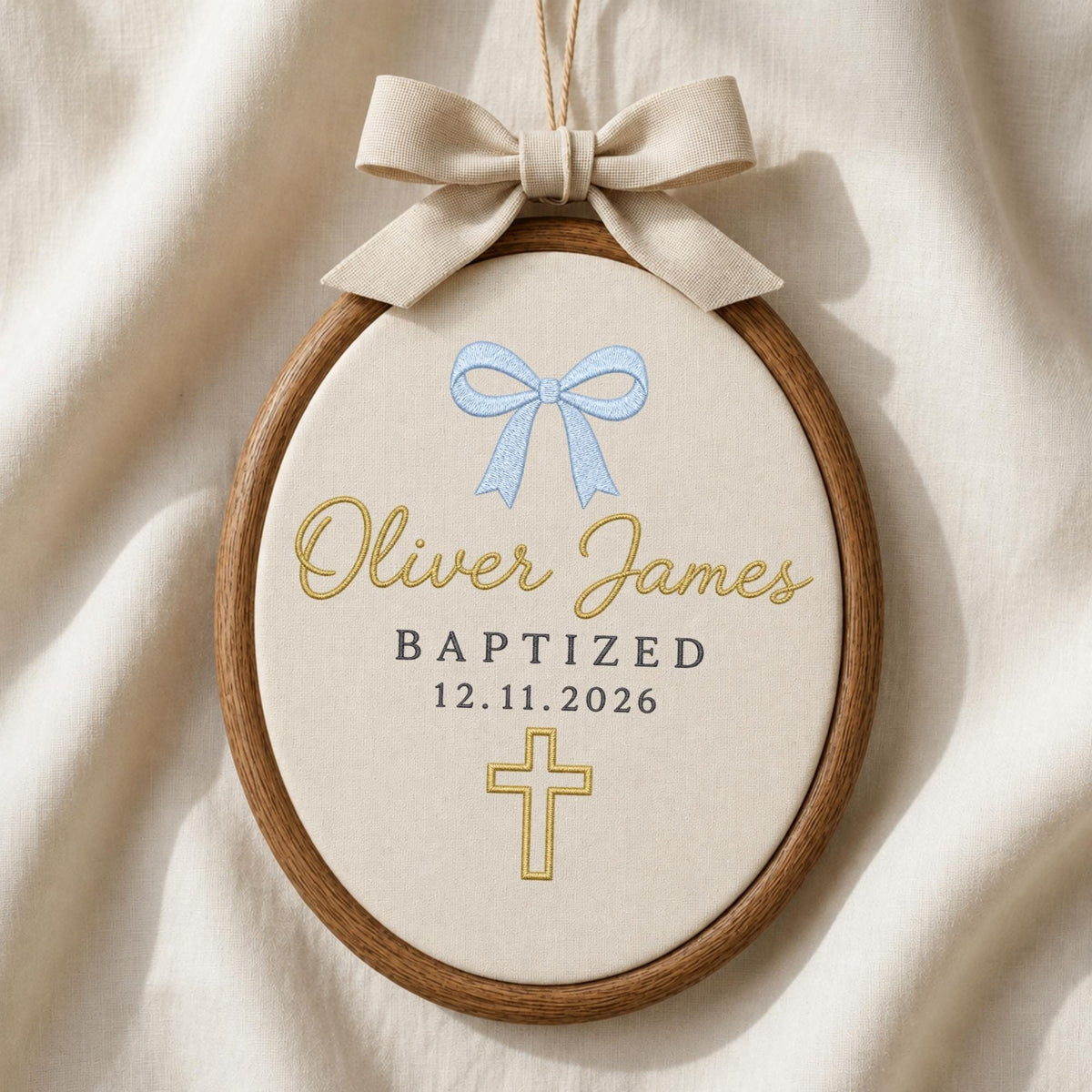 May include: An oval wooden embroidery hoop containing cream fabric with a beige fabric bow at the top. The embroidery features a light blue bow, the name Oliver James in gold script, the text BAPTIZED 12.11.2026 in black, and a gold cross.