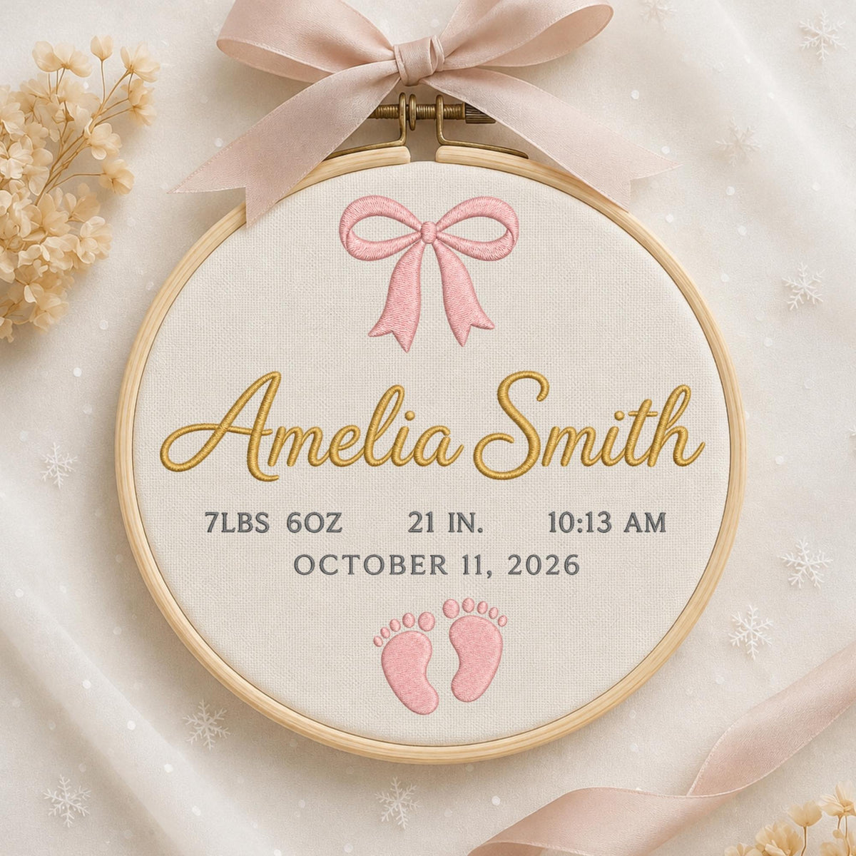 May include: A circular wooden embroidery hoop containing cream fabric with a pink embroidered bow, the name Amelia Smith in gold, and gray text stating 7lbs 6oz, 21 in., 10:13 am, and October 11, 2026, with pink baby footprints and a pink ribbon.
