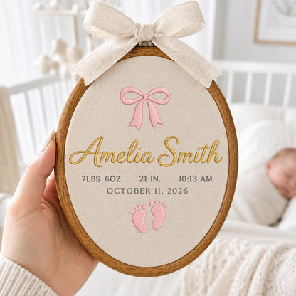 May include: An oval wooden embroidery hoop containing beige fabric with a cream-colored ribbon bow at the top. The embroidery features a pink bow, the name Amelia Smith in gold, and the text 7LBS 6OZ, 21 IN., 10:13 AM, OCTOBER 11, 2026, and pink baby footprints.