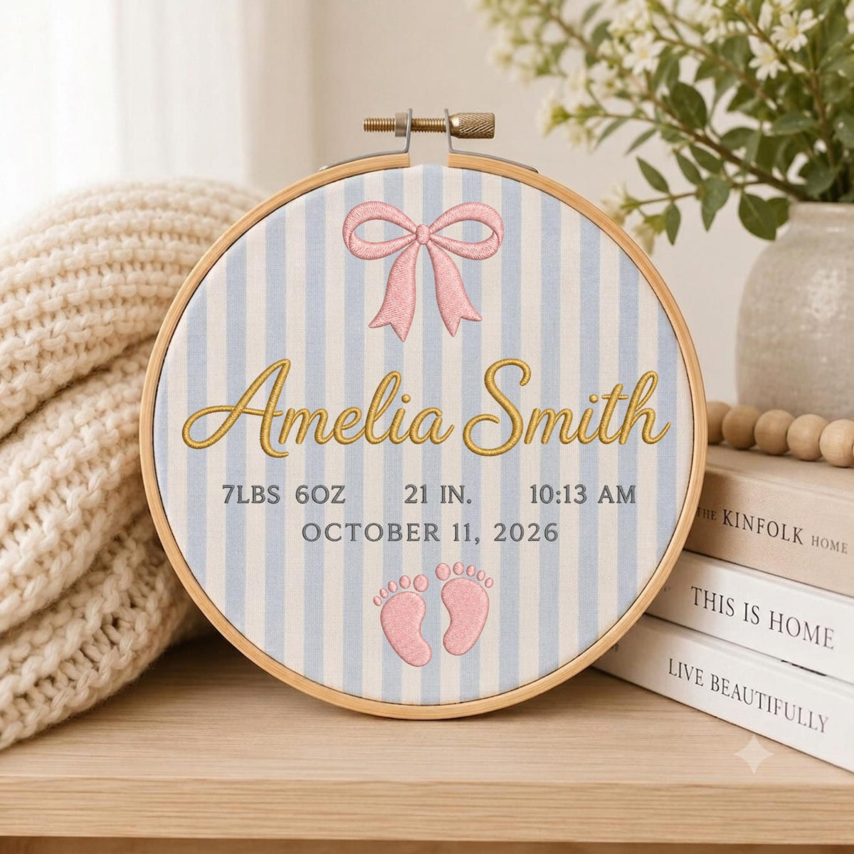 May include: A circular wooden embroidery hoop framing fabric with light blue and white vertical stripes. It features a pink bow, the name Amelia Smith in gold script, and birth details: 7lbs 6oz, 21 in., 10:13 am, October 11, 2026, and pink baby footprints.